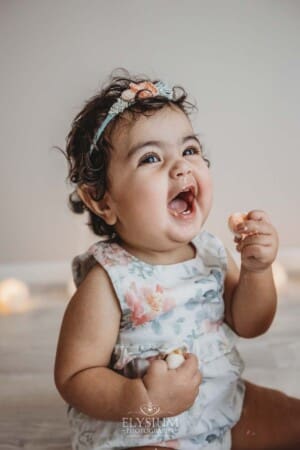 A baby girl smiles as she holds cake in her hand during a cake smash session