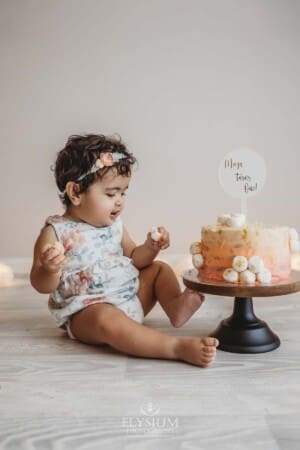 A baby girl sits next to a cake stand with a peach coloured cake for her first birthday