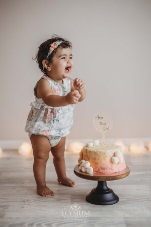 A baby girl giggles as she stands next to her first birthday cake and gets ready to smash it.