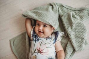 A baby girl dressed in a floral romper lays on her back and smiles peeking out from under a green blanket