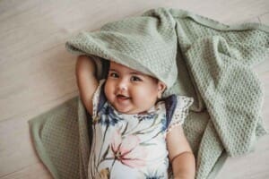 A baby girl dressed in a floral romper plays peek-a-boo with a soft green blanket