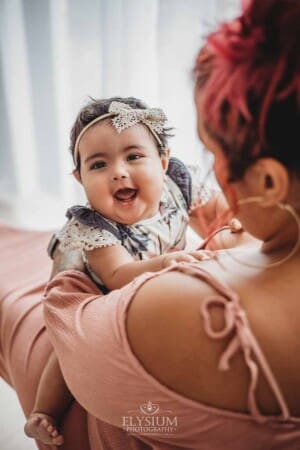 A baby sits in her mothers lap and smiles for a photo next to a bright studio window