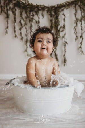 A baby girl splashes water in a bathtub prop and giggles after her cake smash