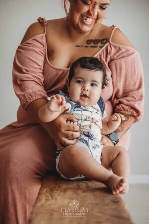 A baby girl sits on a timber bench with her mother and smiles for a photo