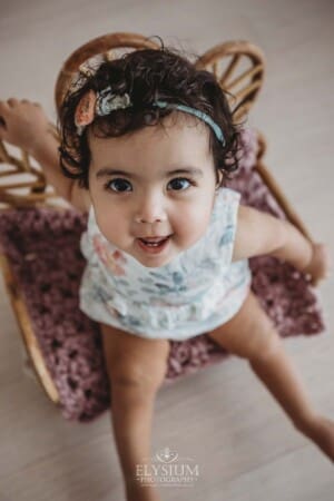 A baby sits on a prop rattan chair wearing a floral romper and smiling for a portrait
