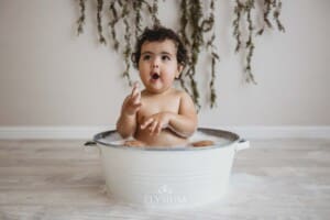 A baby girl sits in a white tub filled with bubbles and smiles as she splashes water