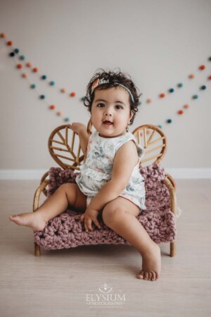 A baby girl in a floral romper sits on a small rattan chair studio prop