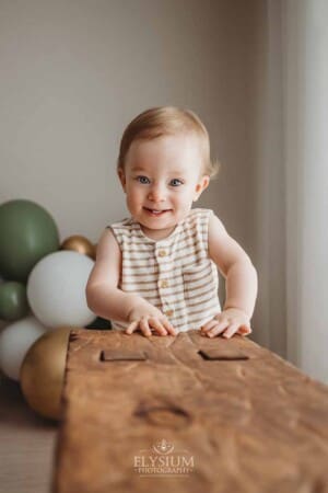 A baby boy holds onto a timber chair in front of his first birthday balloons