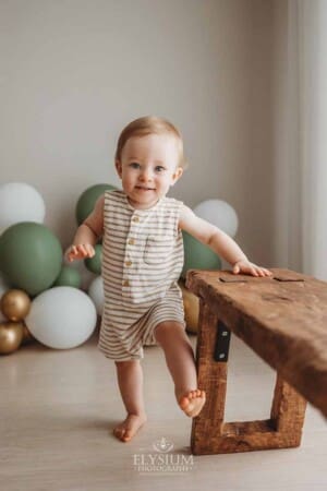 A little boy stands in front of a balloon garland smiles as he leans on a timber bench