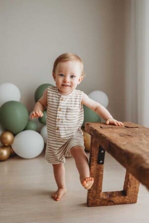 A baby strands next to a timber chair with a balloon garland in the background for his first birthday