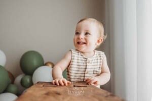 A baby smiles as he balances against a wood bench in a bright studio
