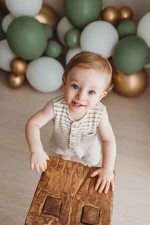 A baby boy standing in front of a timber bench with a balloon garland behind celebrating his first birthday