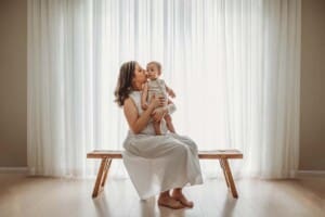 A mother cuddles her baby boy while sitting on a wooden bench in front of a large window