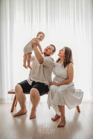 Parents sit close on a wooden bench and hold their smiling baby boy above them