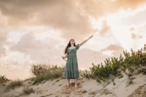 A girl in a green dress stands on a sand dune at sunrise and waves a bubble wand releasing bubbles into the sky