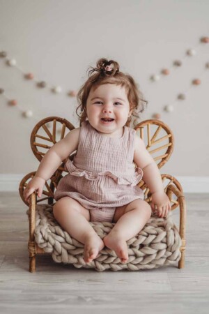 A baby girl in a soft pink romper sits on a small rattan chair in a bright studio