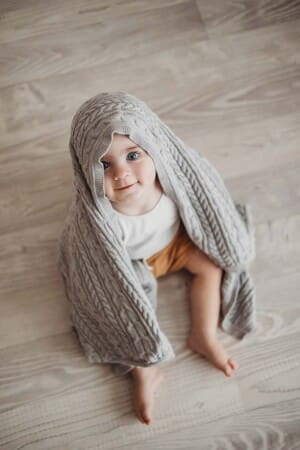 A baby boy peeks out from under a grey knit blanket as he sits on a white wood floor in a studio