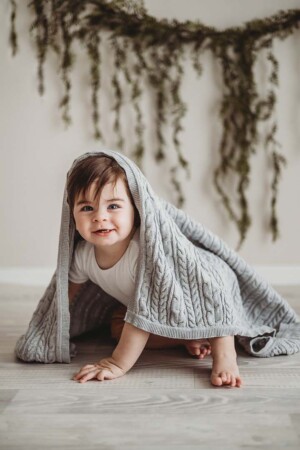 A smiley baby boy crawls along a wood floor with a grey blanket over him for a milestone session