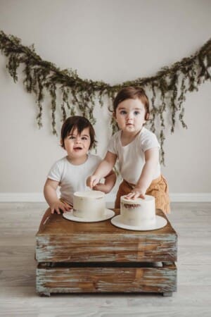 Twin babies stand beside a wood crate about to smash into their matching first birthday cakes