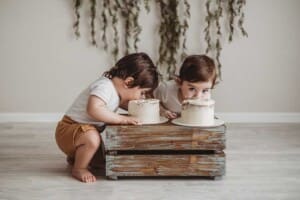 Twin boys sit beside a crate and bite into their first birthday cakes in a studio