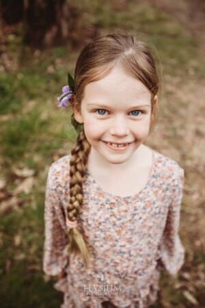 A little girl wearing a floral dress smiles with a purple flower in her hair