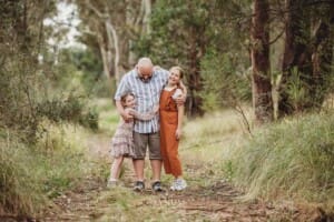 A father hugs his daughters as they stand on a trail in a grassy field