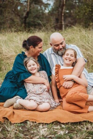 Parents cuddle their daughters as they all sit hugging on a blanket in a field