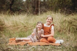 Two girls sit on a blanket in a long grassy field and hug