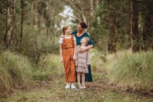 A mother looks lovingly at her two girls as she hugs them in a grassy field