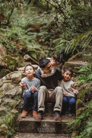 A father sits surrounded by his three kids on sandstone steps