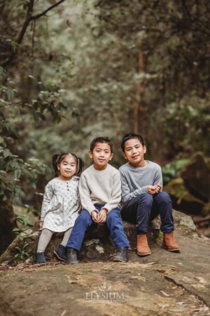 Three kids sit on a large stone surrounded by trees at Lennox Bridge