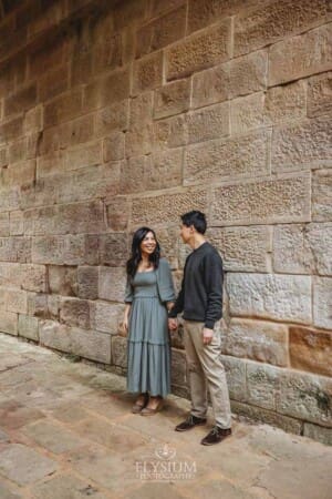 A couple look at each other as they hold hands and stand against a sandstone wall