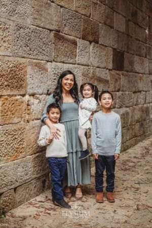 A mother hugs her three children as they stand next to an old sandstone wall
