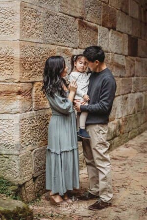 Parents stand against a sandstone wall and hug their little girl between them