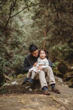A dad holds his little girl in his lap as they sit on a huge rock surrounded by trees