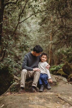 A father sits next to his son hugging him as they sit on a large rock