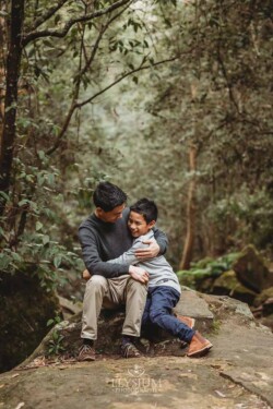 A boy hugs his dad as they sit on a large rock surrounded by trees
