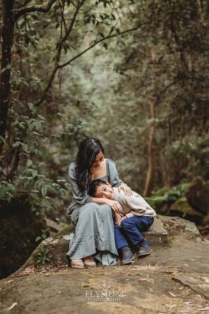 A little boy rests his head in his mothers lap as they sit on a large rock amongst trees