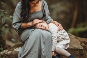 A little girl rests her head in her mothers lap as they sit on a rock surrounded by trees