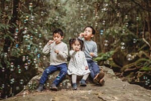 Three siblings sit on a huge rock surrounded by trees and blow bubbles