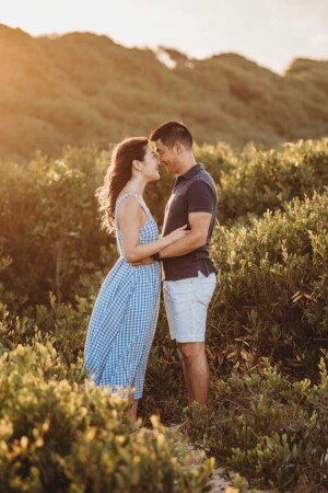 A couple stand in front of a beach dune covered in greenery and cuddle each other at sunset