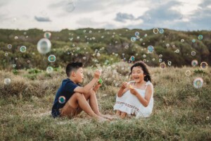 Two children have fun playing with bubbles while sitting in grass on a sand dune at Maroubra