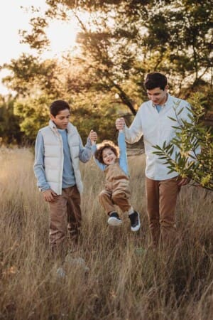 Three siblings have fun in a grassy field lifting their baby brother between them as he giggles