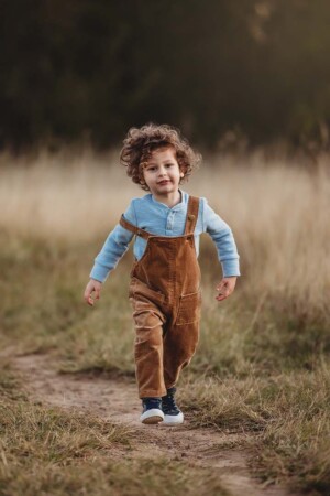 A baby boy wearing overalls and a blue shirt skips along a dirt track through long grass at sunset