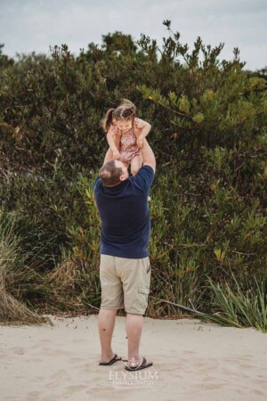 A dad hoists his little girl above him as they stand on a beach