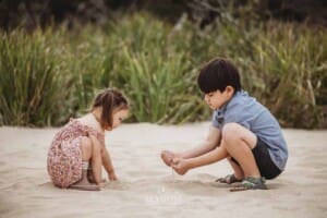 Two children play with the sand as they crouch together on a beach