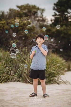 A little boys stands on a beach with trees behind him as he blows bubbles