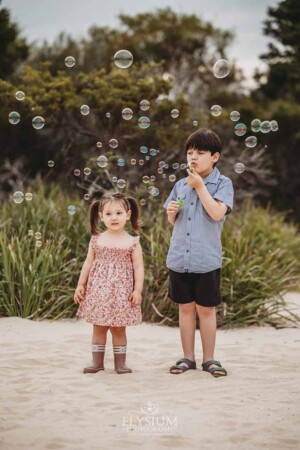 Two children stand on a beach surrounded by greenery and blow bubbles