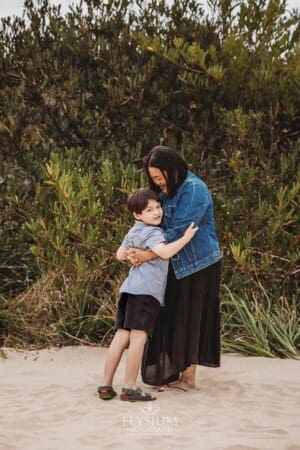 A mother hugs her son as they stand on beach sand surrounded by trees