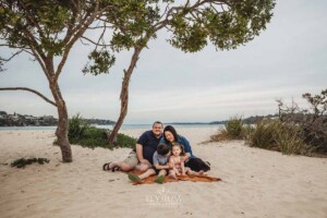 A family sit on a rug at the beach and hug each other under a tree